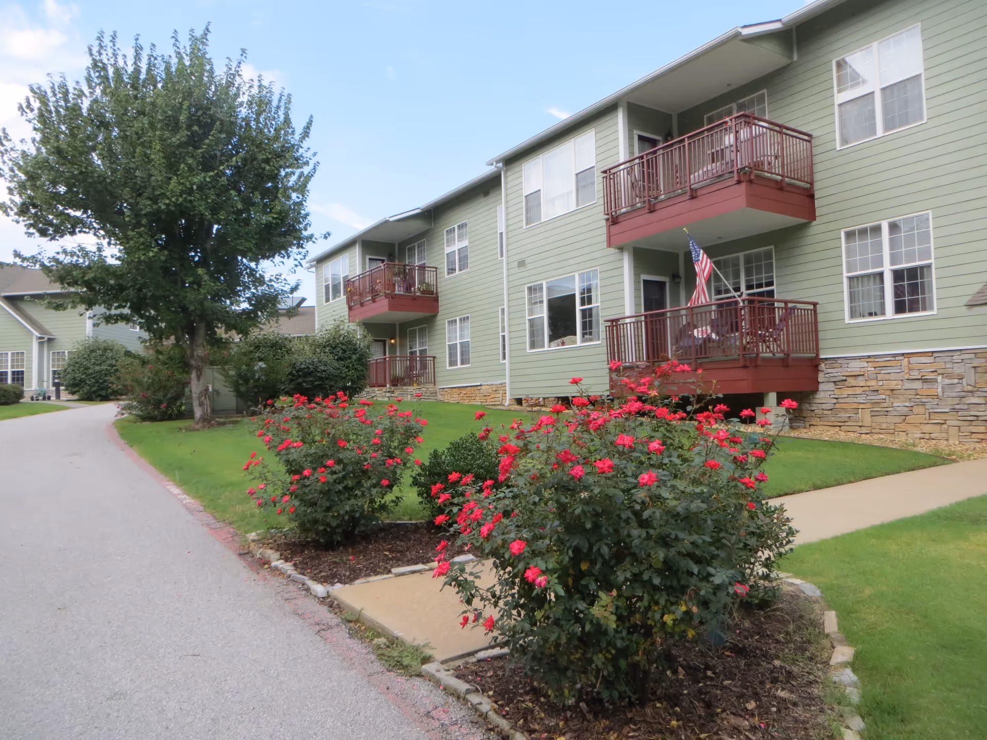 Two-story green apartment building with red balconies, landscaped lawn and rose bushes along a driveway.