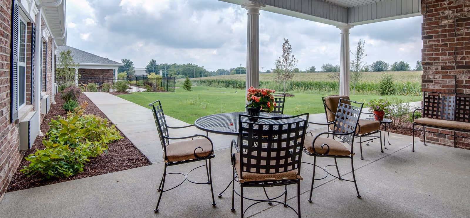 Covered outdoor patio area with a round metal table and four metal chairs with cushions. There are potted flowers on the table and additional seating along the brick wall. The patio overlooks a grassy field with trees and a cloudy sky.