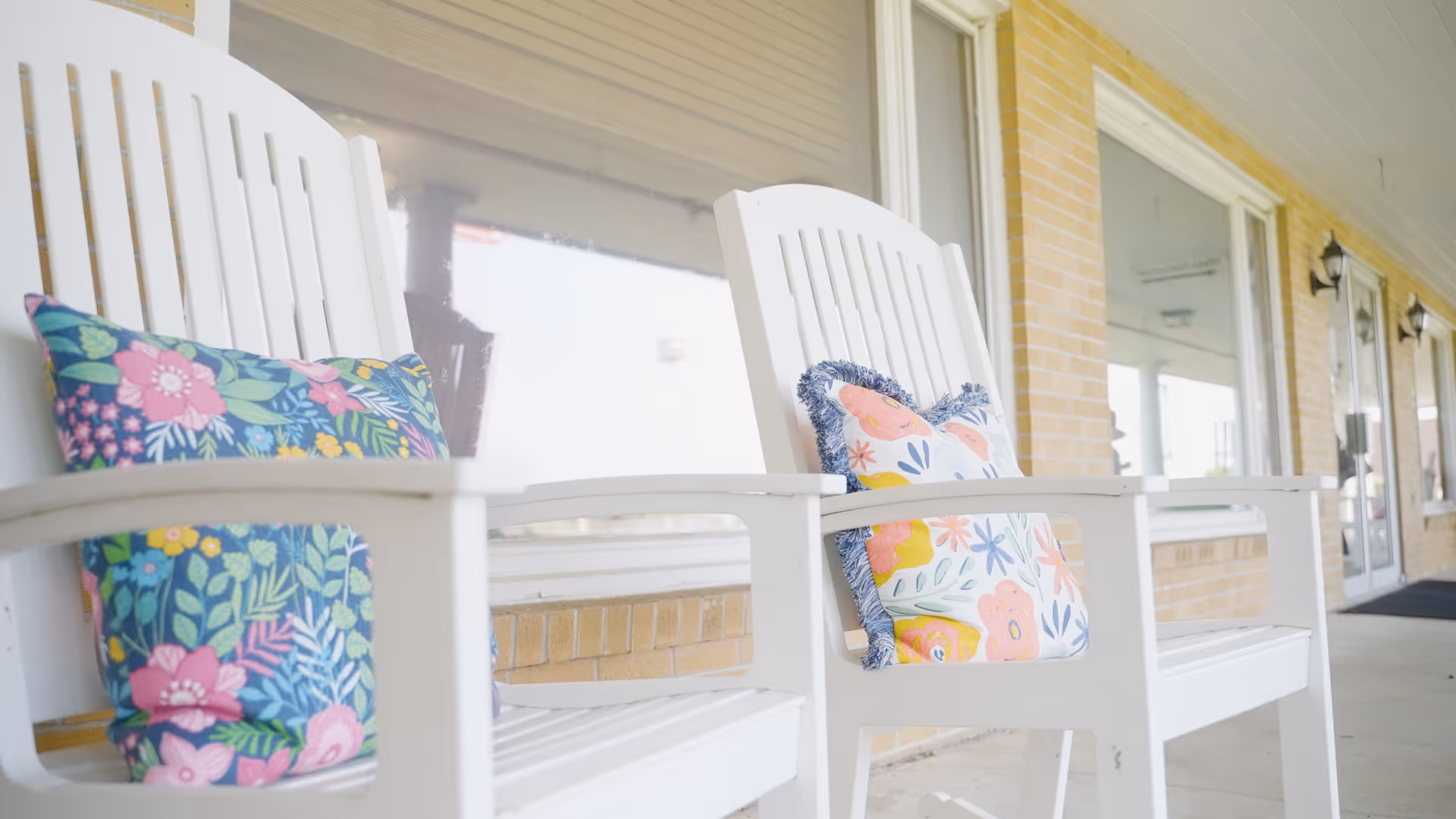 Two white wooden chairs with colorful floral cushions placed on a covered porch outside a building with large windows and brick walls.