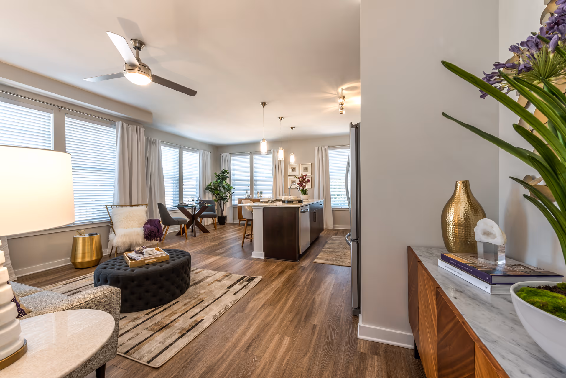 Bright and modern open-concept living space with large windows covered by white blinds and curtains, featuring a ceiling fan, a cozy seating area with a gray sofa, a tufted black ottoman, a small round side table with a lamp, a dining area with a small round table and chairs, and a kitchen island with pendant lights above. Decorative plants and vases add a touch of greenery and elegance.