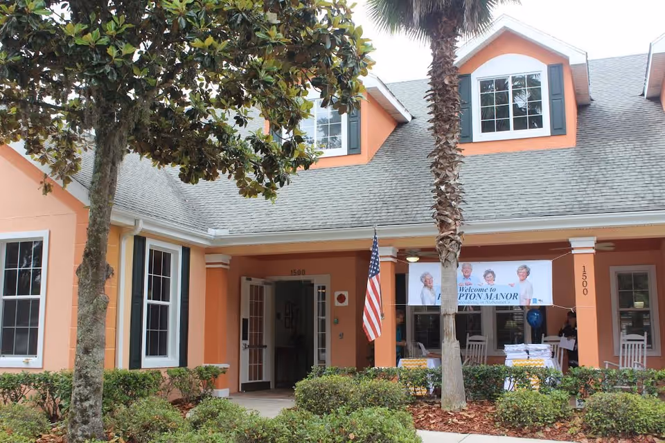 Front exterior view of Hampton Manor Assisted Living at 24th Road, showing a peach-colored building with white trim, large windows, an American flag, and a banner welcoming visitors. There are trees and bushes in the foreground.