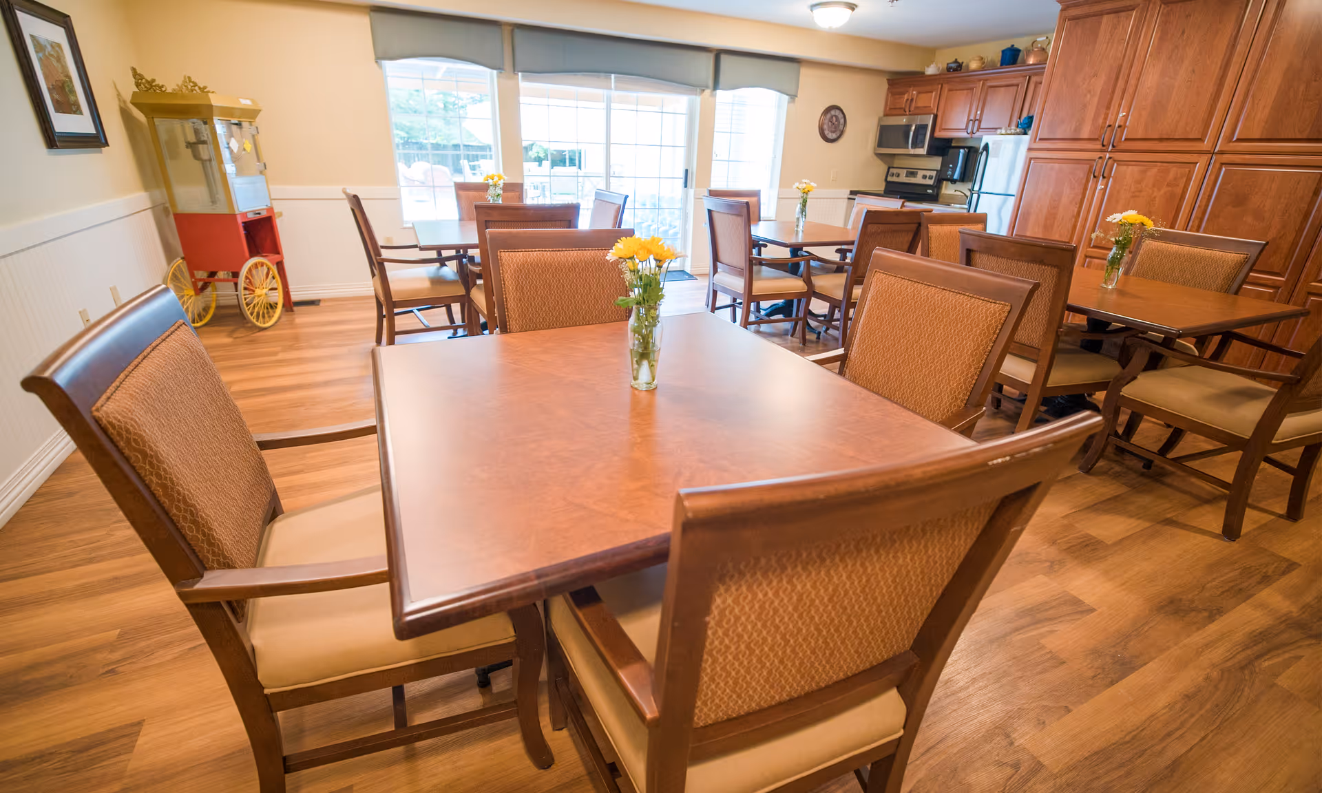 A bright dining room with wooden tables and chairs, each table decorated with a small vase of yellow flowers. The room has large windows with blinds, wooden flooring, and a popcorn machine in the corner. There is a kitchen area with wooden cabinets and stainless steel appliances in the background.