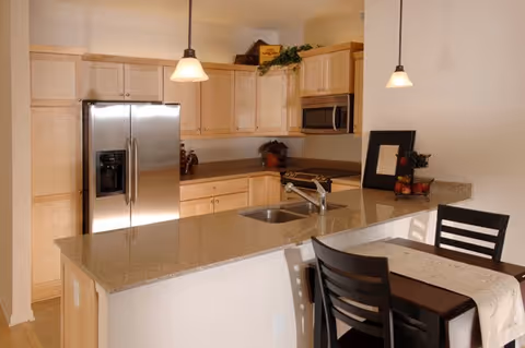 Modern kitchen with light wood cabinets, stainless steel refrigerator, built-in microwave, granite countertops, a sink with a faucet on the island, two pendant lights hanging from the ceiling, and a small dining table with two black chairs and a table runner.