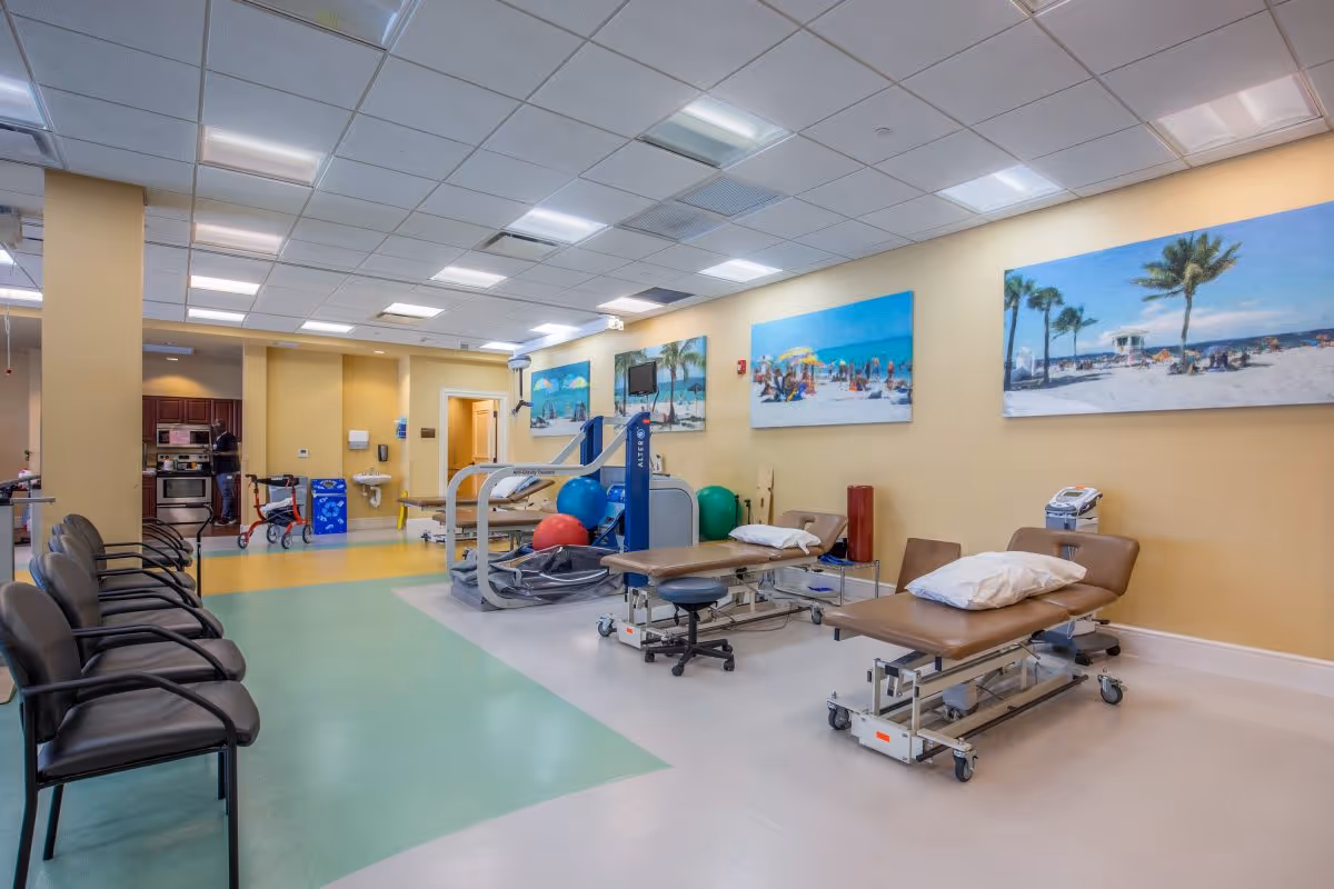 A spacious therapy or rehabilitation room with several brown treatment tables equipped with pillows, exercise equipment including large exercise balls, and a row of black chairs along one wall. The walls are painted yellow and decorated with large beach-themed paintings. In the background, there is a kitchenette area and a person standing near the counter.