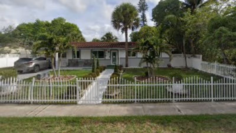 Front exterior view of a single-story building with a red-tiled roof, white walls, and a green door, surrounded by a white picket fence and landscaped with palm trees and grass. A car is parked on the driveway to the left.