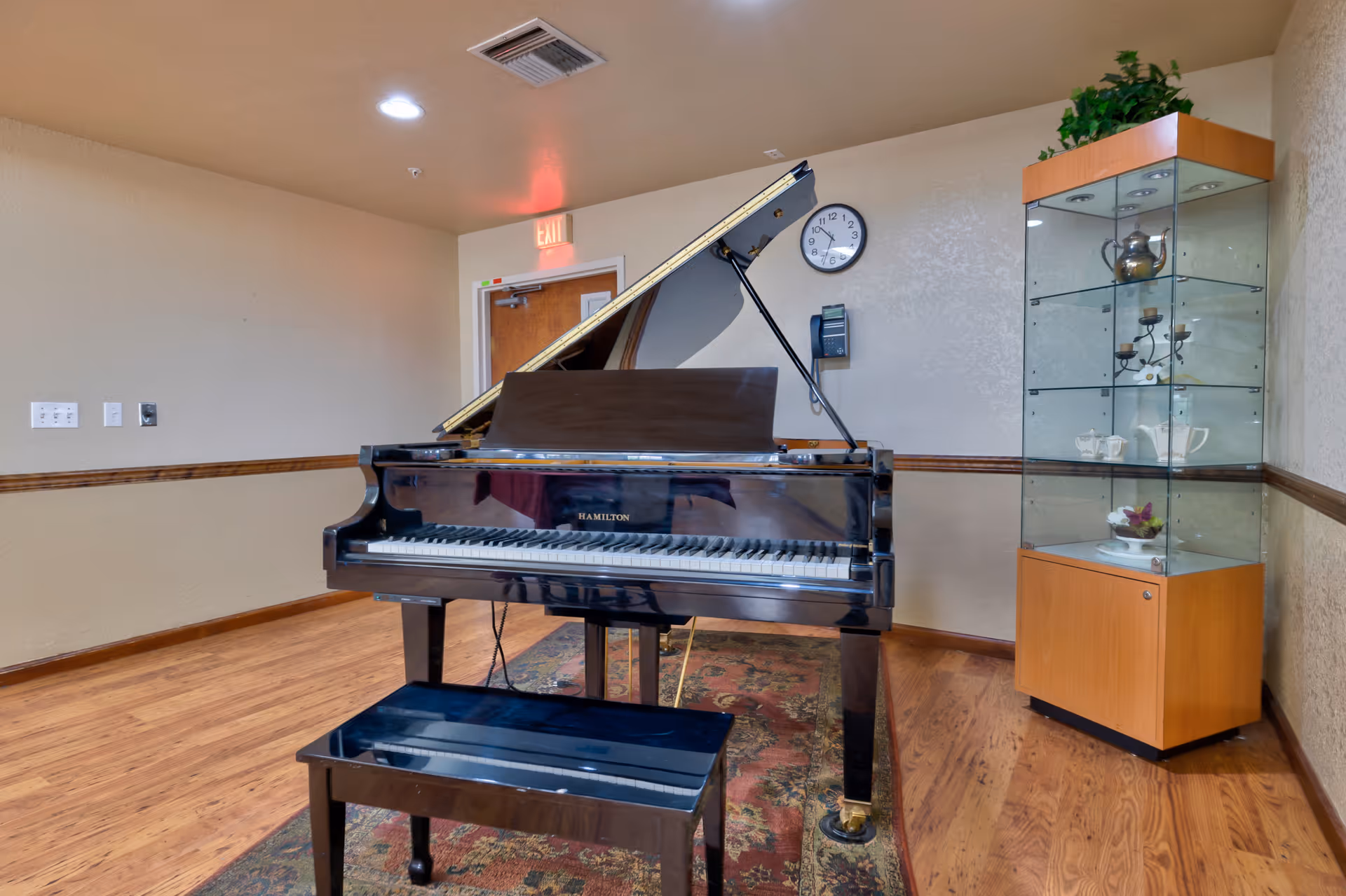 A black grand piano and bench sit on a rug in a communal room with wood floors, a glass display cabinet, a wall clock, and an exit door.