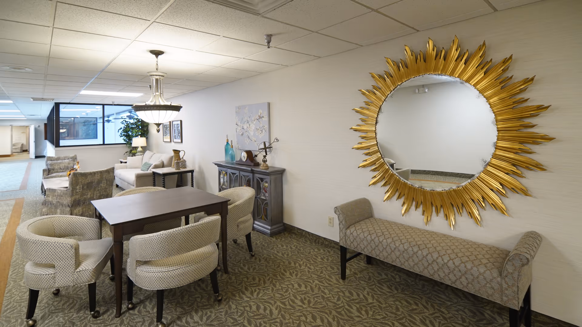 A cozy senior living community interior with a small wooden table surrounded by four cushioned chairs. To the right, there is a decorative bench beneath a large round mirror with a golden sunburst frame. In the background, there is a sitting area with a sofa, armchairs, a side table with a lamp, and decorative items on a cabinet against the wall. The space has carpeted flooring and a drop ceiling with fluorescent lighting.