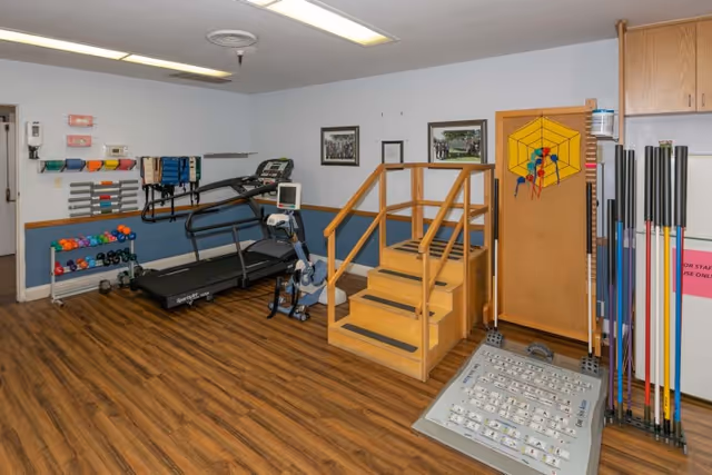A fitness and therapy room with wooden flooring, featuring a treadmill, exercise bike, various colorful dumbbells on racks, resistance bands hanging on the wall, a wooden staircase with handrails, and a pegboard game on a stand. The walls are white with a blue lower half, and framed pictures hang on the wall.