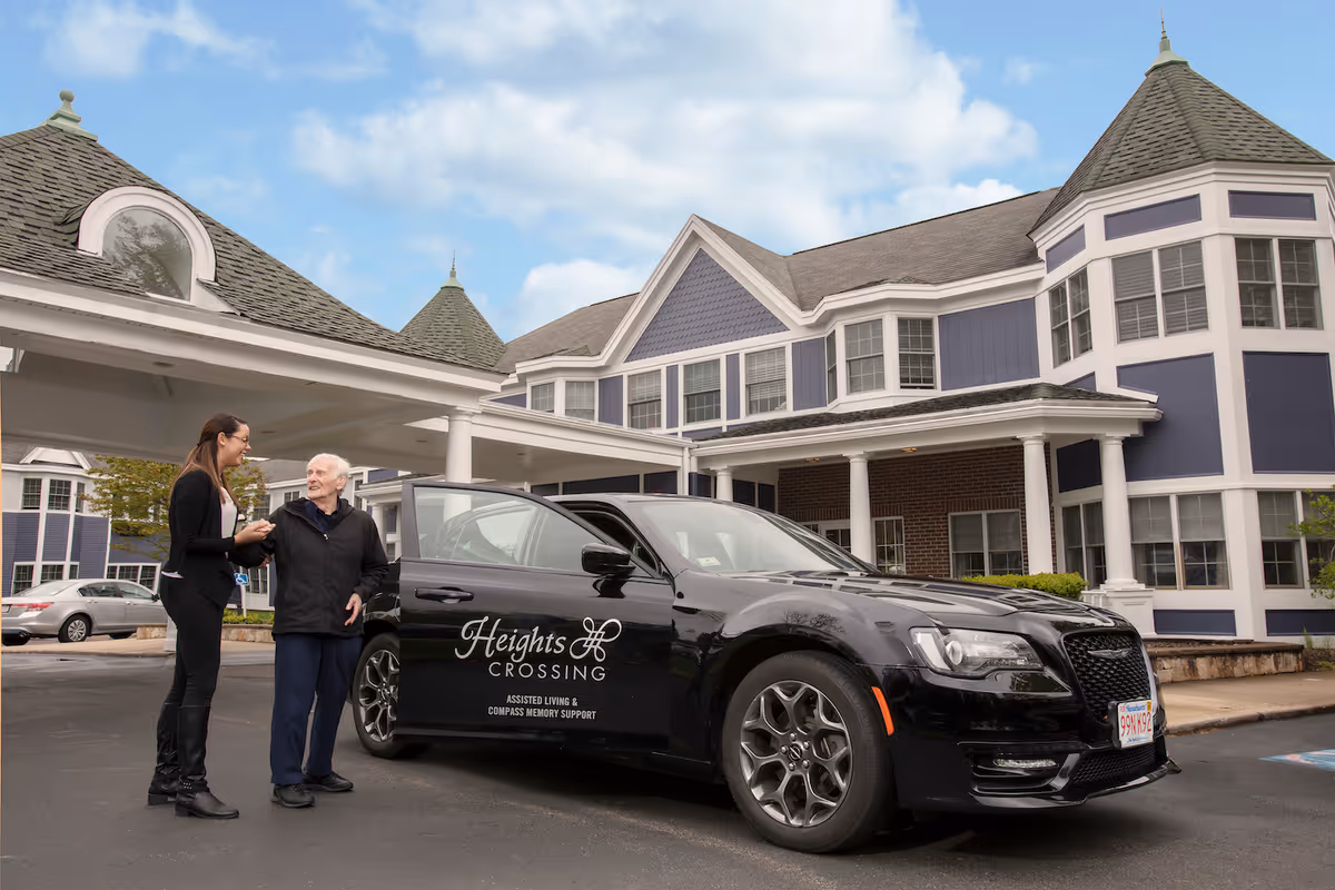 An elderly man and a younger woman standing next to a black car with the logo 'Heights Crossing Assisted Living & Compass Memory Support' on the door, in front of a large, multi-story building with blue and white exterior and multiple windows under a partly cloudy sky.