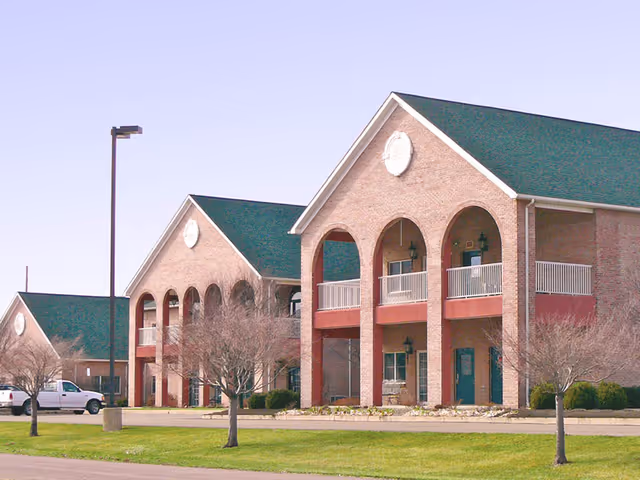 Exterior view of a senior living facility named Vintage Knolls featuring multiple two-story brick buildings with green roofs, arched balconies, and leafless trees in front. A white pickup truck is parked near the buildings under a clear sky.
