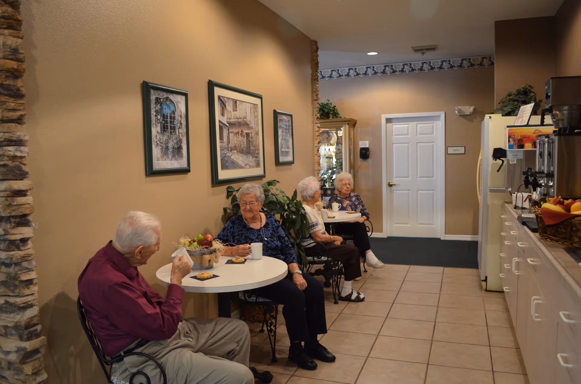 Four elderly residents sit at small round tables in a cozy senior living dining area with wall art and a coffee station.