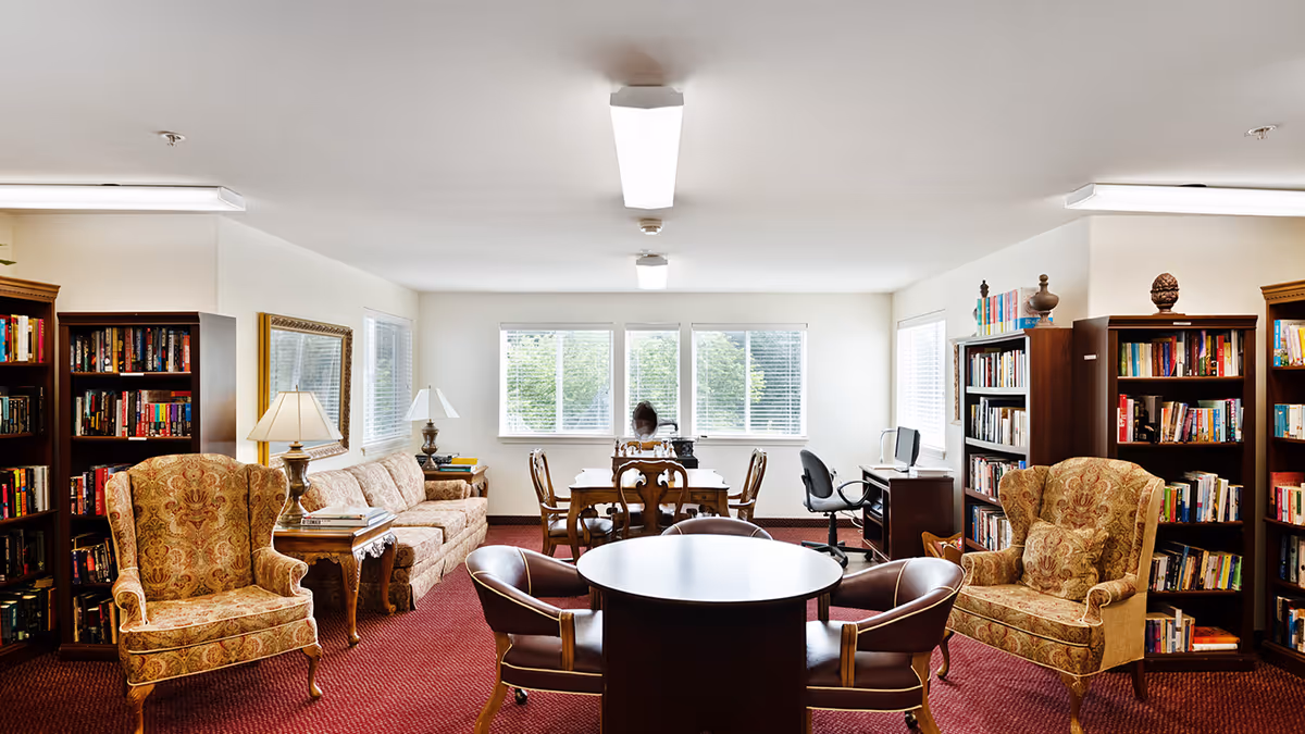 A well-lit common room with large windows, featuring two patterned armchairs, a sofa, multiple bookshelves filled with books, a round table with four chairs, a rectangular table with chairs, and a desk with a computer chair. The room has a red carpet and white walls.