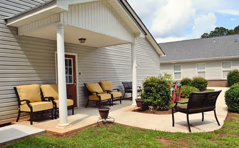 Outdoor patio area at Brookside Commerce with cushioned chairs and benches arranged on a concrete walkway next to a building with beige siding and a red door under a covered porch. There are green bushes and grass surrounding the patio area under a partly cloudy sky.