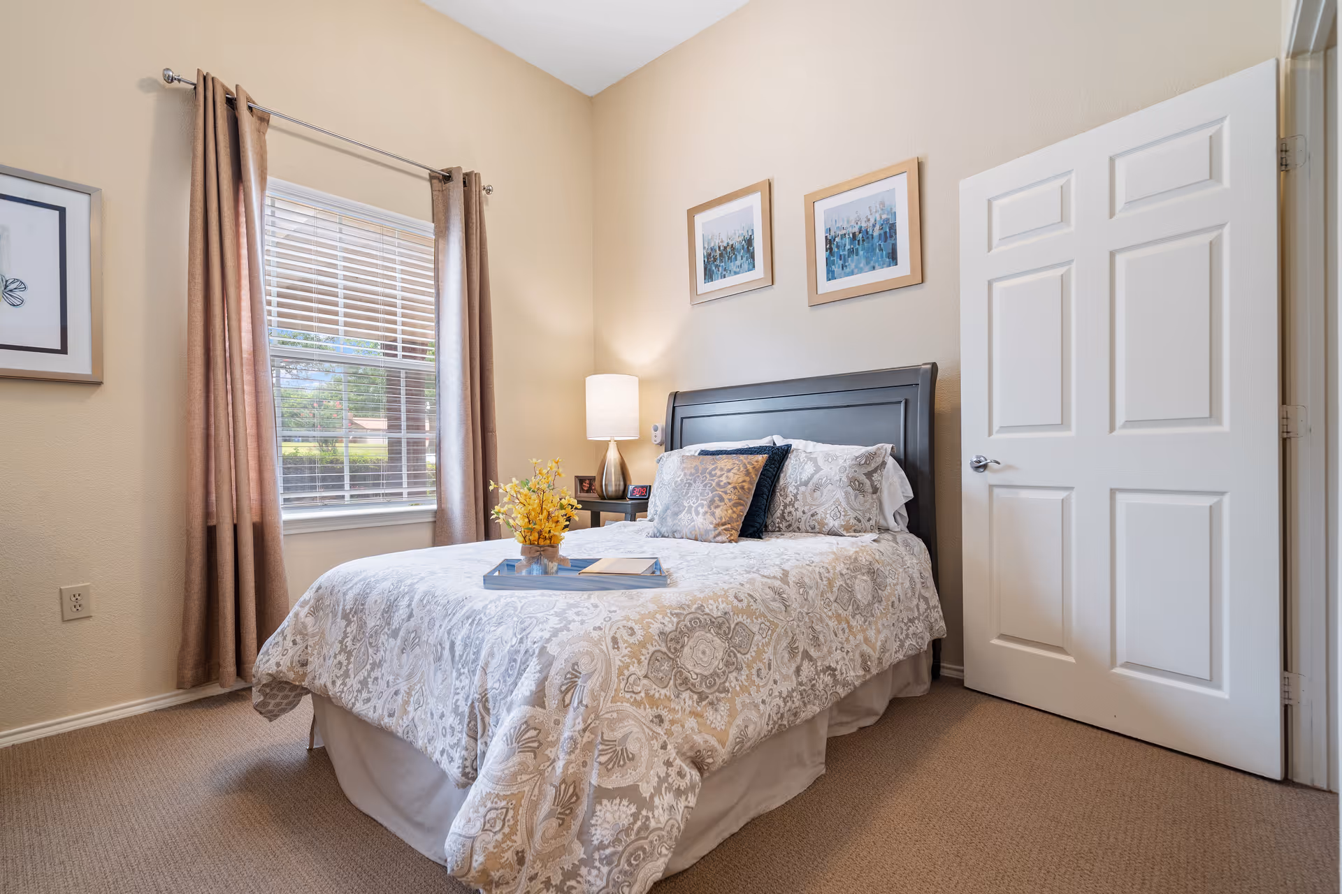 A cozy bedroom with a neatly made bed featuring patterned bedding and decorative pillows. There is a bedside table with a lamp and a small clock. The room has beige walls, a window with brown curtains, and three framed pictures hanging above the bed. The door to the room is open.