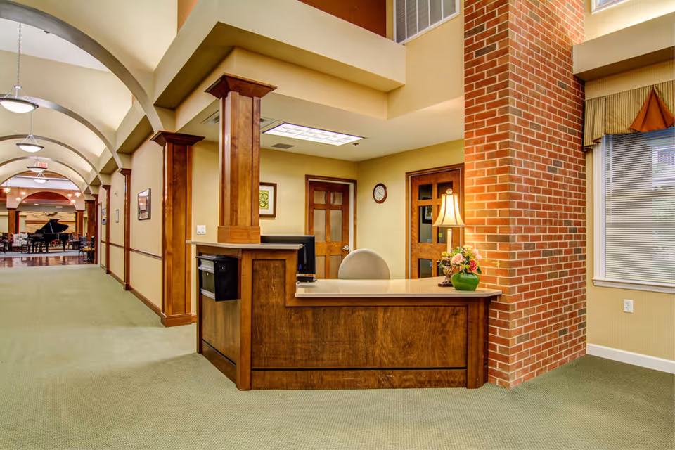 Reception desk in a senior living facility lobby with wood trim, a brick column, arched hallway and a lounge area with a piano visible down the corridor.
