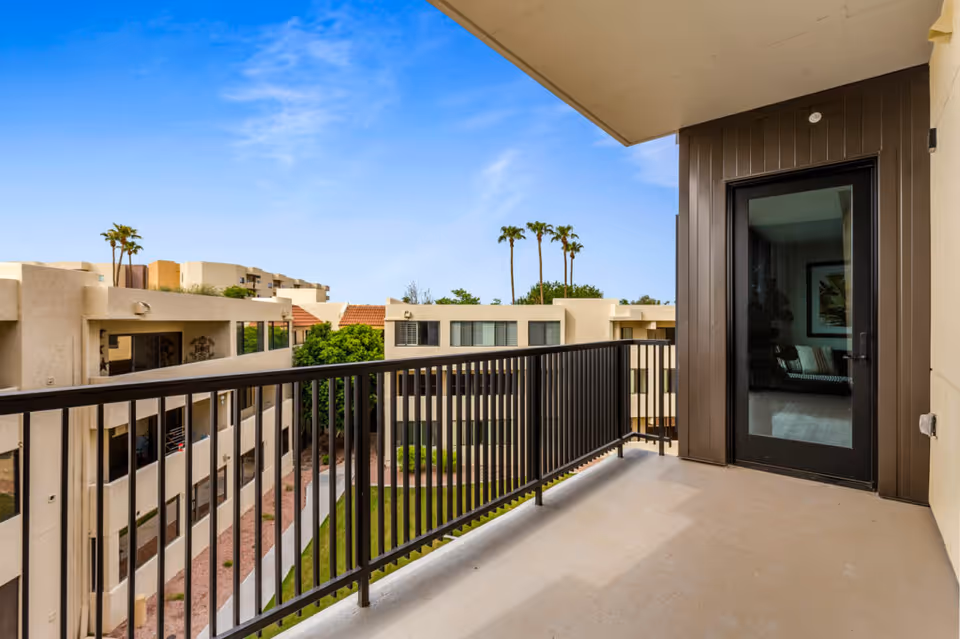 Balcony with a metal railing overlooking a courtyard and adjacent apartment buildings under a blue sky.