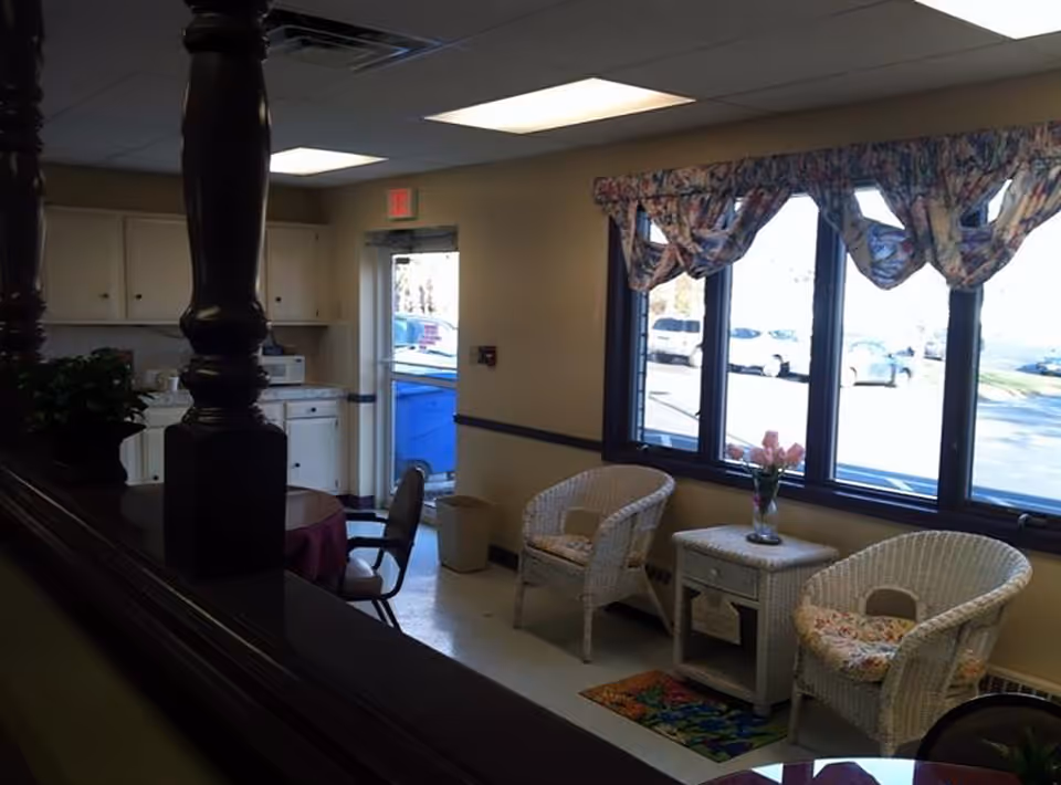 Interior view of a common area with two white wicker chairs with floral cushions, a small white side table with a vase of flowers, large windows with floral valances, and a door leading outside. There is a table with chairs and kitchen cabinets in the background.