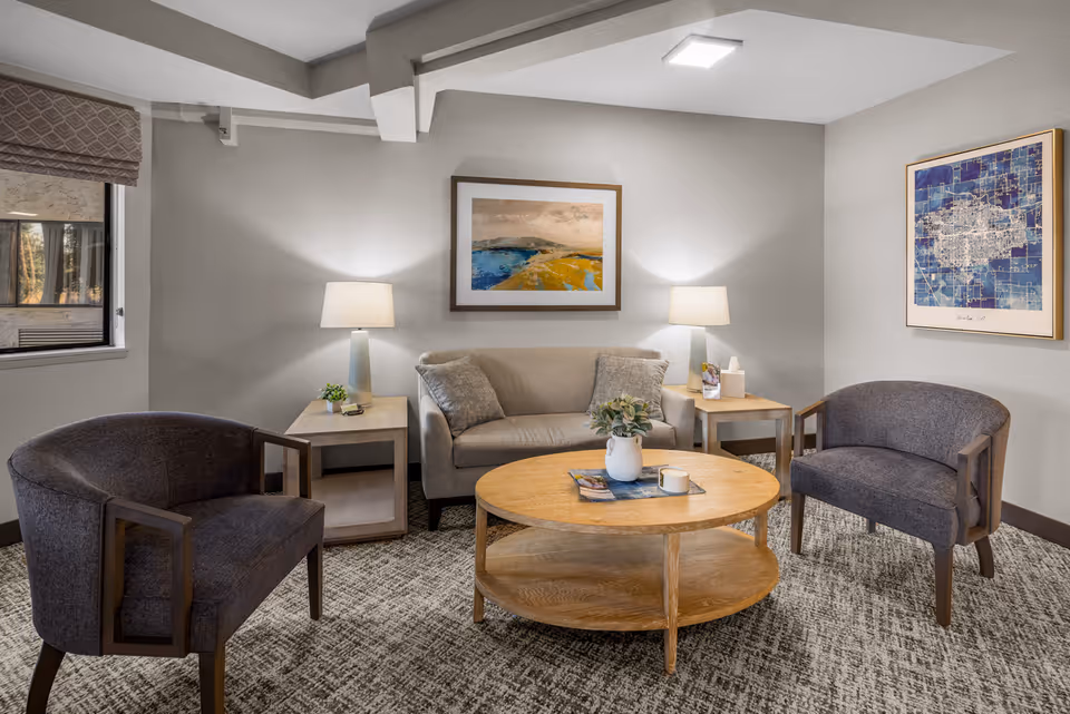 A cozy sitting area in a senior living community featuring a beige loveseat with two gray pillows, two dark gray armchairs, a round wooden coffee table with a white vase and a candle, two wooden side tables each with a lamp, and two framed artworks on the light gray walls.