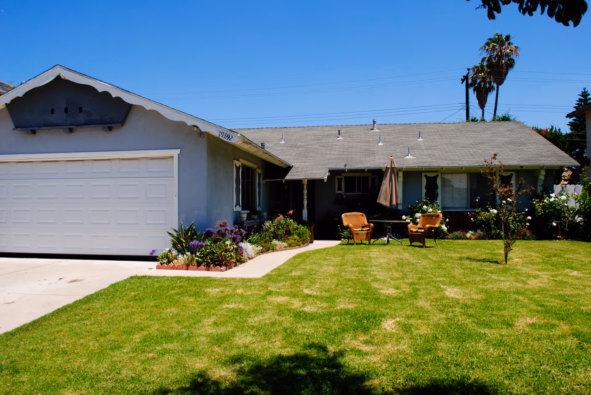 Single-story house with a gray roof and light blue exterior walls, a white garage door, a well-maintained green lawn, flower beds along the front, two wicker chairs and a table with a closed umbrella on the lawn, and palm trees in the background under a clear blue sky.