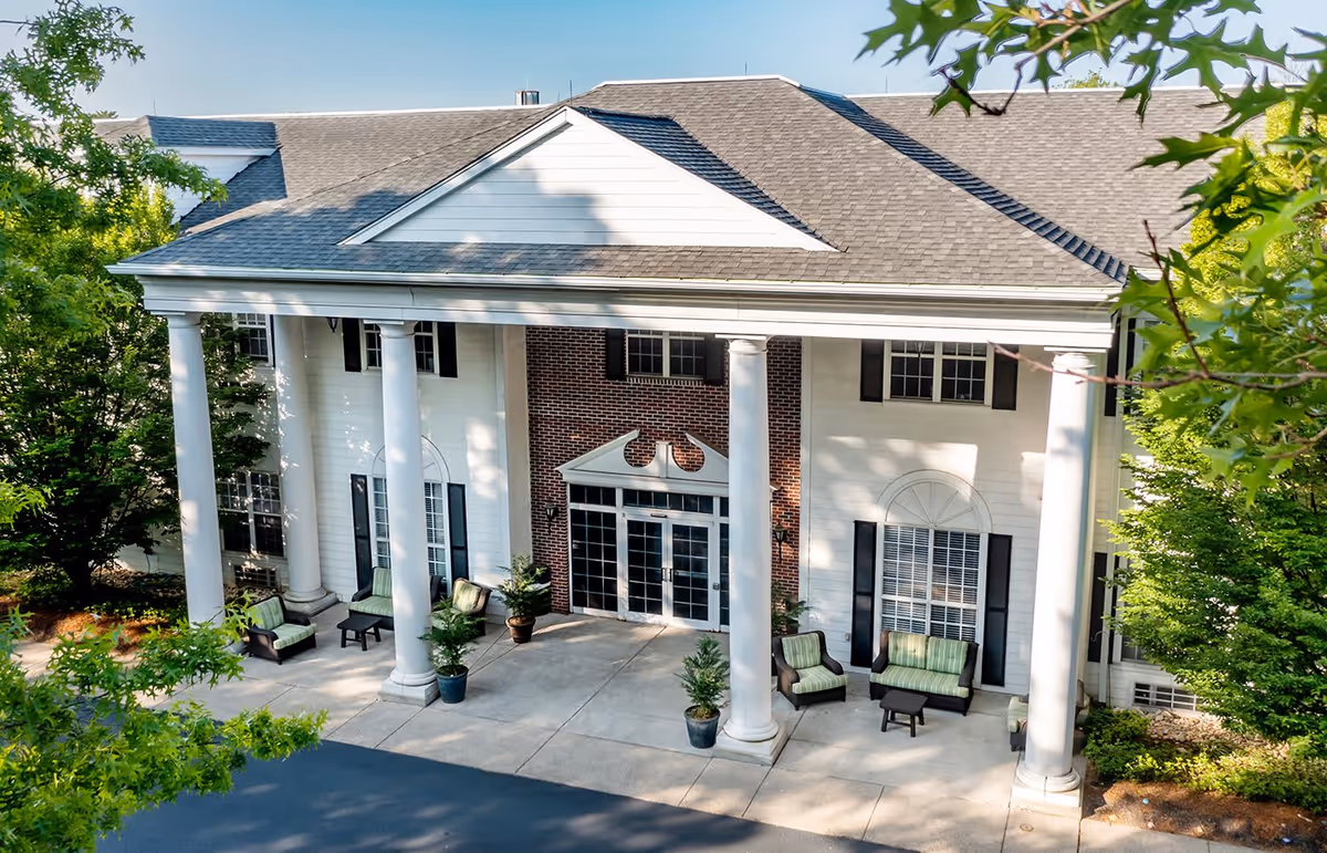 Front exterior view of a senior living facility named Blue Bell Place featuring large white columns, a brick facade, multiple windows with black shutters, and outdoor seating with green cushions surrounded by trees.