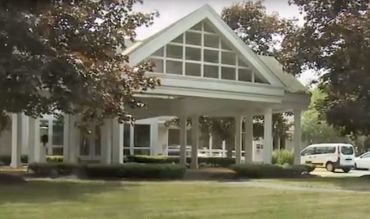 Covered front entrance of a senior living building with white columns, a triangular gable of windows, landscaping, and a parked van.