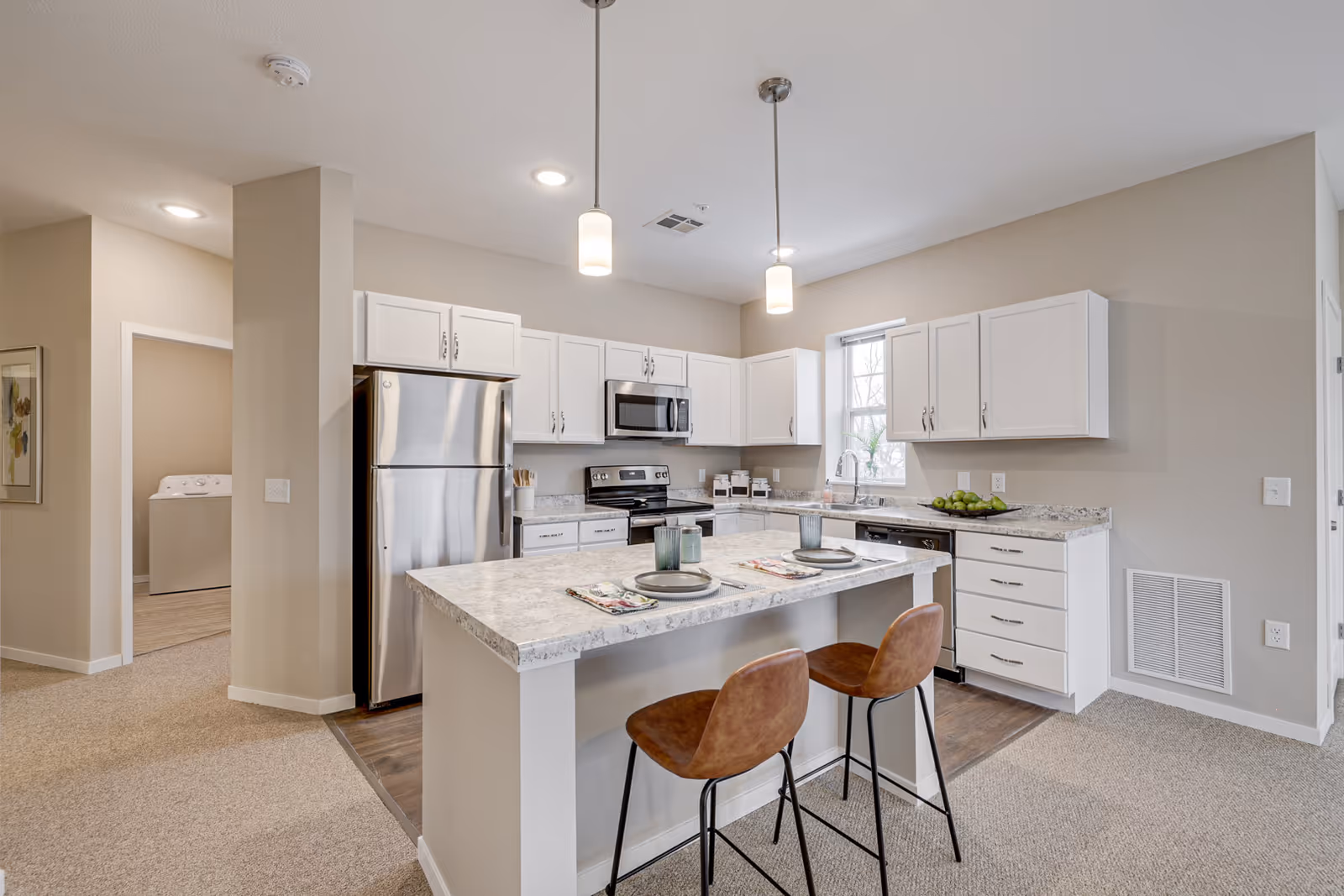 Modern kitchen with white cabinets, stainless steel refrigerator, stove, microwave, and dishwasher. A kitchen island with a marble countertop has two brown bar stools and place settings for two. Two pendant lights hang above the island, and a window above the sink lets in natural light. The kitchen floor is wood, transitioning to carpet in the adjacent area.