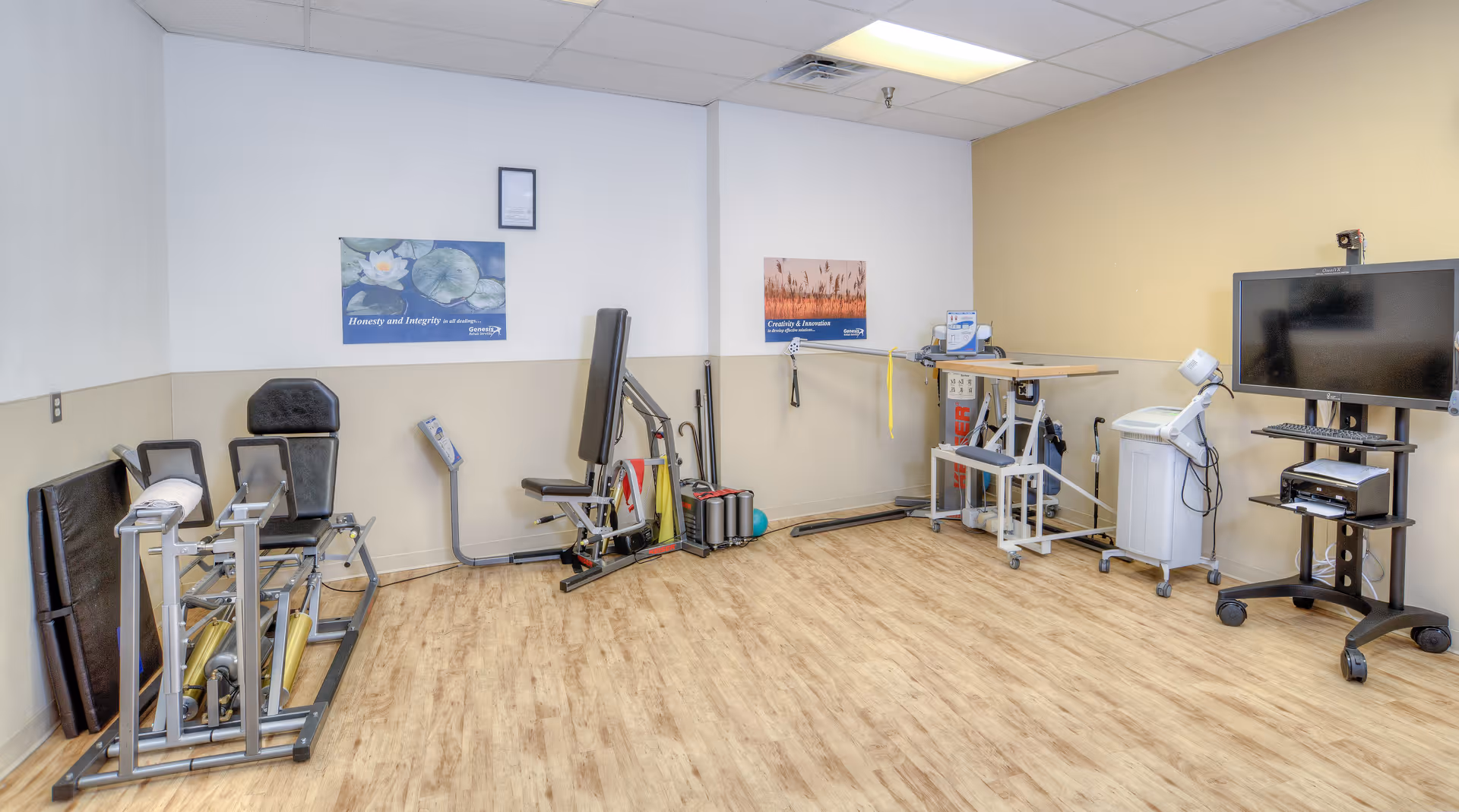 A rehabilitation therapy room with exercise equipment including a leg press machine, a seated exercise machine, and various physical therapy tools. The room has light wood flooring, white and beige walls, two motivational posters, and a large TV on a stand with attached medical devices.
