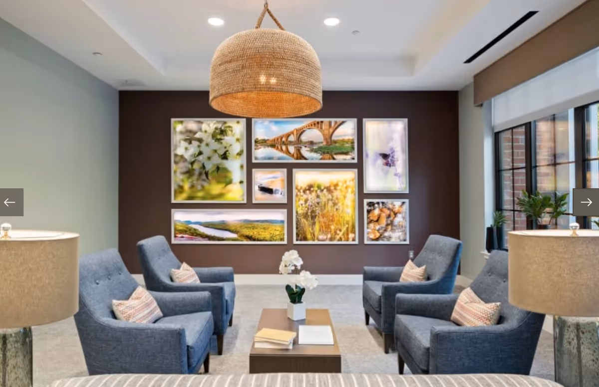 Bright sitting area with four blue armchairs around a coffee table facing a gallery wall of framed photos under a woven pendant light.