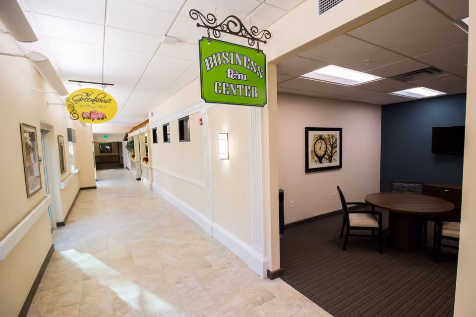 Interior hallway of a senior living facility with beige walls and tiled floor. On the right, there is a business center room with a round table and chairs, a wall clock, and a TV mounted on a dark blue accent wall. Hanging signs indicate the Business Center and The Greenhouse on Village Drive.