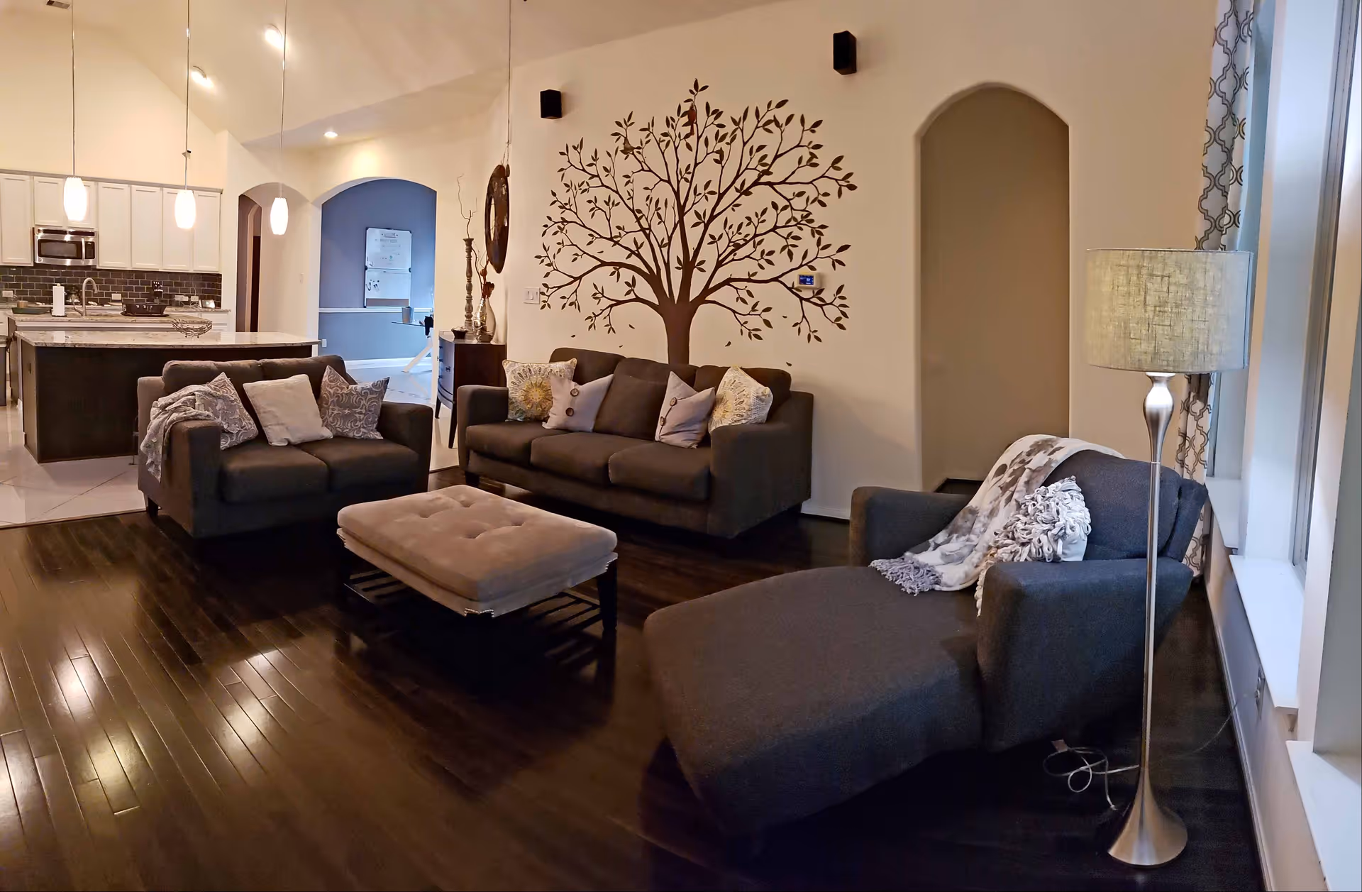 A cozy living room with dark hardwood floors, featuring a dark gray sofa, loveseat, and chaise lounge adorned with decorative pillows and throws. A tufted ottoman sits in the center. The wall behind the sofa has a large tree decal. To the left, an open kitchen area with white cabinets, a kitchen island, and pendant lights is visible. A floor lamp stands next to the chaise lounge by a window with patterned curtains.
