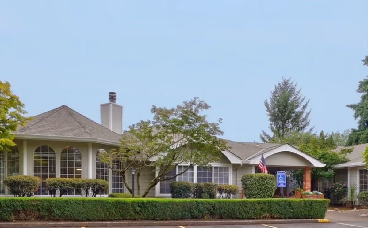Exterior view of a single-story building with large windows, a covered entrance with an American flag, surrounded by neatly trimmed bushes and trees under a clear blue sky.