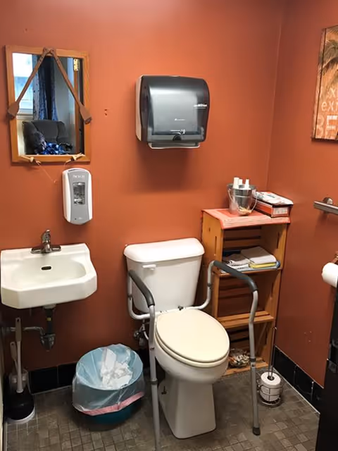 A small bathroom with orange walls featuring a white toilet equipped with safety rails, a white sink with a faucet, a wall-mounted soap dispenser, a paper towel dispenser, a small wooden shelf holding various items, a trash can with a liner, and a mirror hanging on the wall.