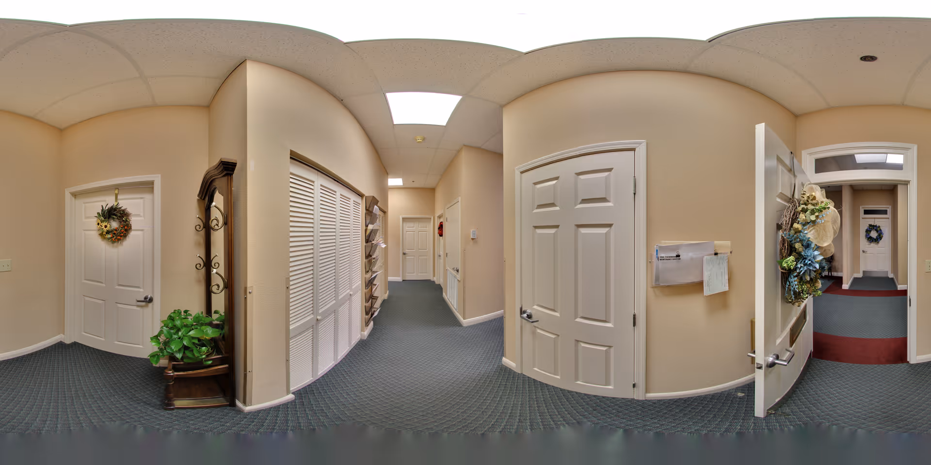 Carpeted interior hallway of a senior living facility with several doors, decorative wreaths, a potted plant, and mail slots on the wall.