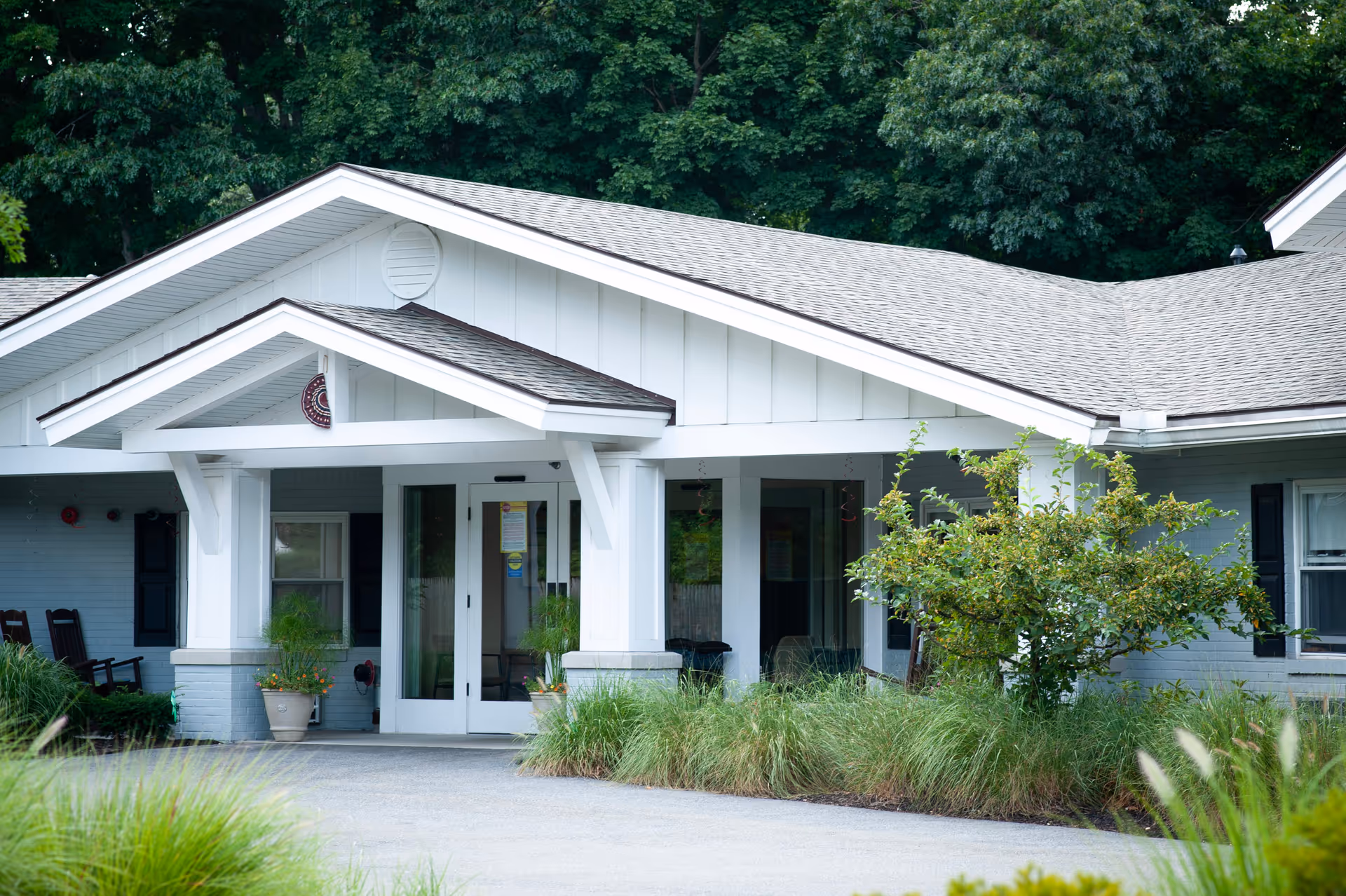 White building front with a covered entry, columns, and surrounding shrubs.