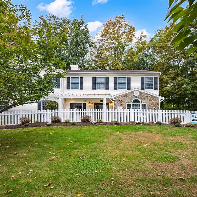 A two-story residential building with white siding and black shutters, featuring a stone accent wall on one side. The building is surrounded by a white picket fence and landscaped with small bushes and a green lawn. Trees with green and autumn-colored leaves are visible in the background under a partly cloudy blue sky.