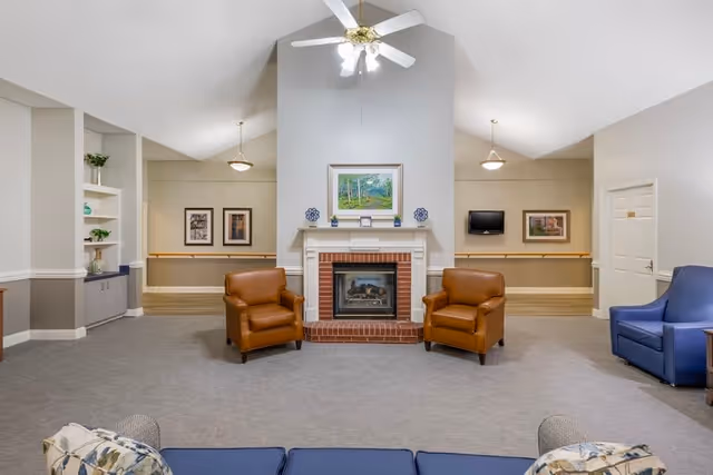 A spacious senior living common area with a high ceiling and ceiling fan. The room features a brick fireplace centered on the far wall, flanked by two brown leather armchairs. To the right, there is a blue armchair and a small wooden side table. The walls are painted light gray with white trim, and there are framed pictures and a small TV mounted on the walls. Built-in shelves with decorative items are visible on the left side.