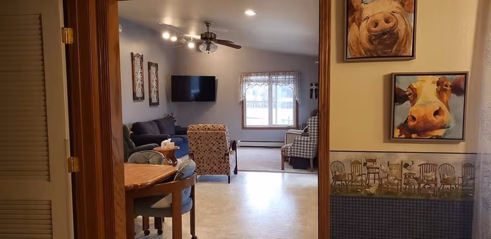 View through a doorway into a living room area with a sofa, armchairs, a wall-mounted TV, and a window with lace curtains. In the foreground, there is a wooden dining table with chairs. The wall on the right has two paintings of cows and a wallpaper border featuring various wooden chairs.