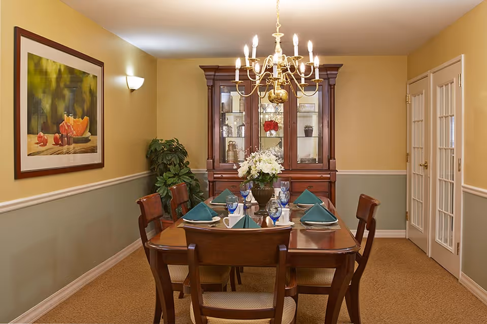A formal dining room with a wooden dining table set for six people with green napkins, blue glasses, and white plates. A chandelier with lit candles hangs above the table. Behind the table is a wooden china cabinet with glass doors displaying decorative items. The walls are painted yellow and green with a framed painting of fruit on the left wall and a potted plant in the corner. There are double French doors on the right side of the room.