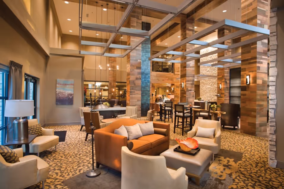 A spacious and well-lit living room area in a senior living facility featuring a brown sofa with gray pillows, beige armchairs, a large ottoman with a decorative bowl, and multiple tables and chairs. The room has high ceilings with modern hanging light fixtures, wooden pillars, and a patterned carpet. Large windows with curtains allow natural light to enter the space.