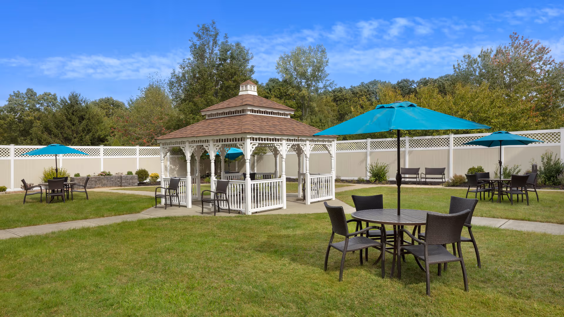 Outdoor garden area with green grass, a white gazebo with a brown roof, several round tables with black chairs, and blue umbrellas. The area is enclosed by a white fence with lattice design on top, and there are trees and shrubs in the background under a blue sky with some clouds.