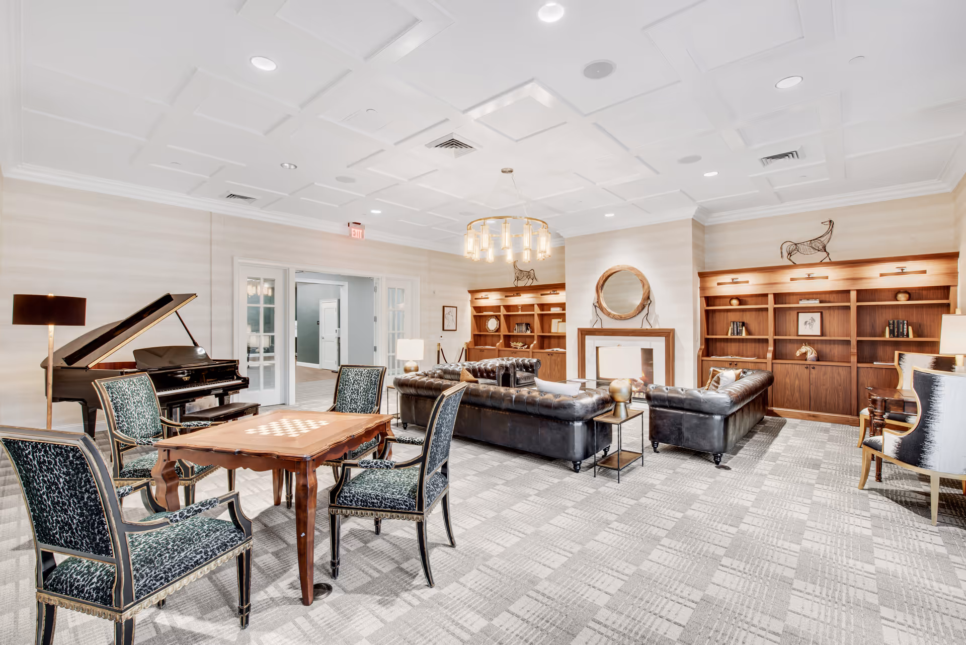 A spacious and elegant living room area with a grand piano on the left, a wooden table with four upholstered chairs in the foreground, and two black leather sofas arranged around a fireplace with a round mirror above it. The room features built-in wooden bookshelves with decorative items, a modern chandelier, and a patterned carpet floor.