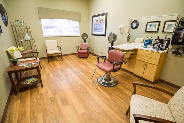 A well-lit room with wooden flooring featuring a salon chair in front of a sink and mirror, two hair dryer chairs against the wall, and several cushioned chairs arranged around the room. A small table with magazines and a flower vase is also visible, along with a window with blinds and framed pictures on the walls.