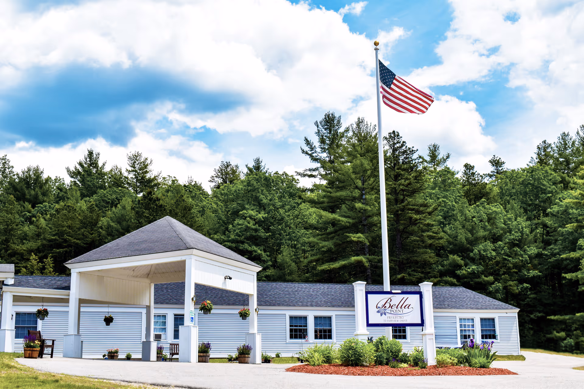 Front exterior of Bella Point Fryeburg showing a single-story building with a covered entrance, landscaping, a sign, and an American flag on a tall pole.