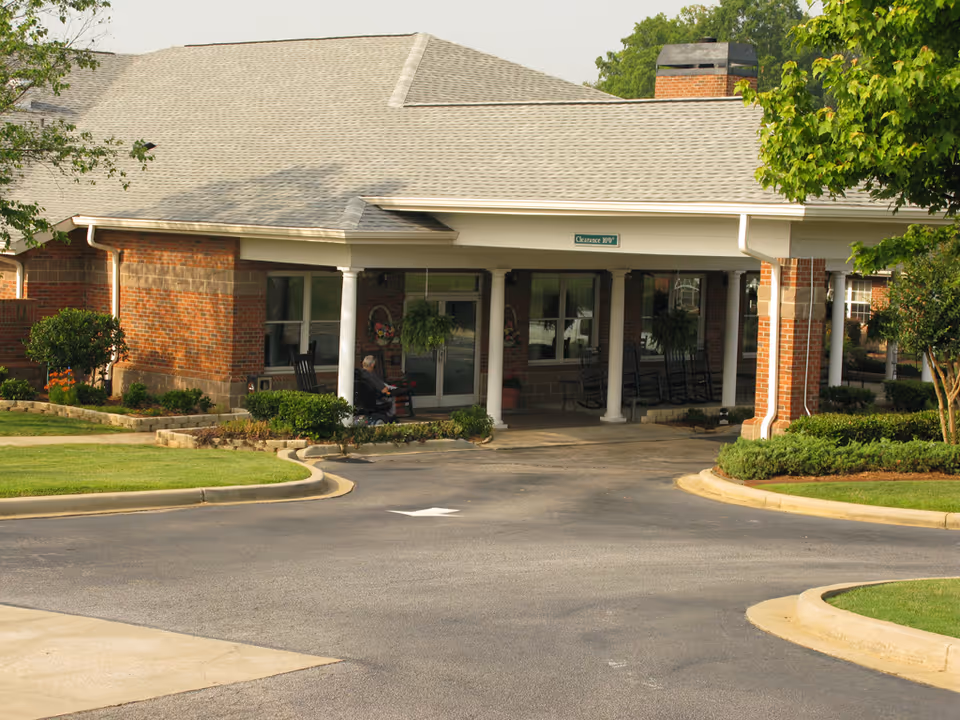Front entrance of a brick building with a covered driveway, white columns, and a gray shingled roof. There are rocking chairs on the porch and greenery including bushes and trees around the building. A person in a wheelchair is near the entrance.
