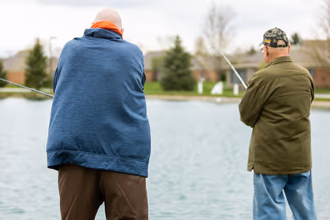 Two elderly men fishing by a pond in an outdoor setting with trees and buildings in the background.