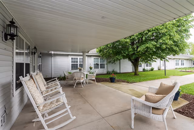 Covered outdoor patio area with white rocking chairs and cushioned wicker chair, a table with chairs in the background, and a large tree with green leaves near a walkway in front of single-story buildings.