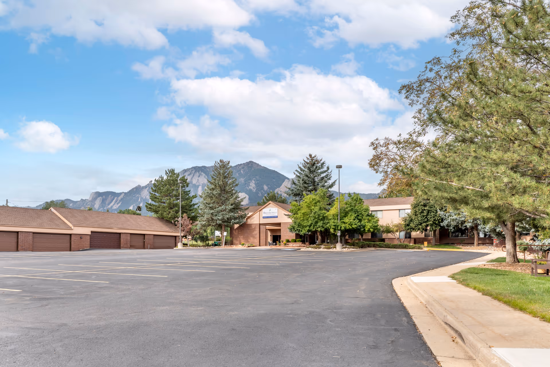 Exterior view of Brookdale Meridian Boulder senior living facility with a large empty parking lot in the foreground, surrounded by trees and mountains in the background under a partly cloudy sky.