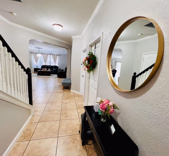 A hallway with beige tiled floor leading to a living room area with black leather sofas and a coffee table. On the right wall of the hallway, there is a round mirror with a gold frame above a black console table decorated with a vase of pink flowers. A wreath hangs on a white door along the hallway. The walls are light gray and the ceiling has a light fixture.
