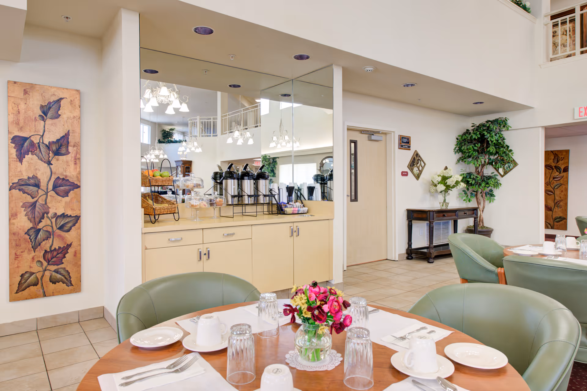 Interior view of a senior living facility dining area with round tables set with plates, cups, utensils, and glasses. Green cushioned chairs surround the tables. A counter with coffee dispensers, baskets of fruit, and pastries is visible against a mirrored wall. Decorative plants and artwork adorn the walls, and a door and hallway are seen in the background.