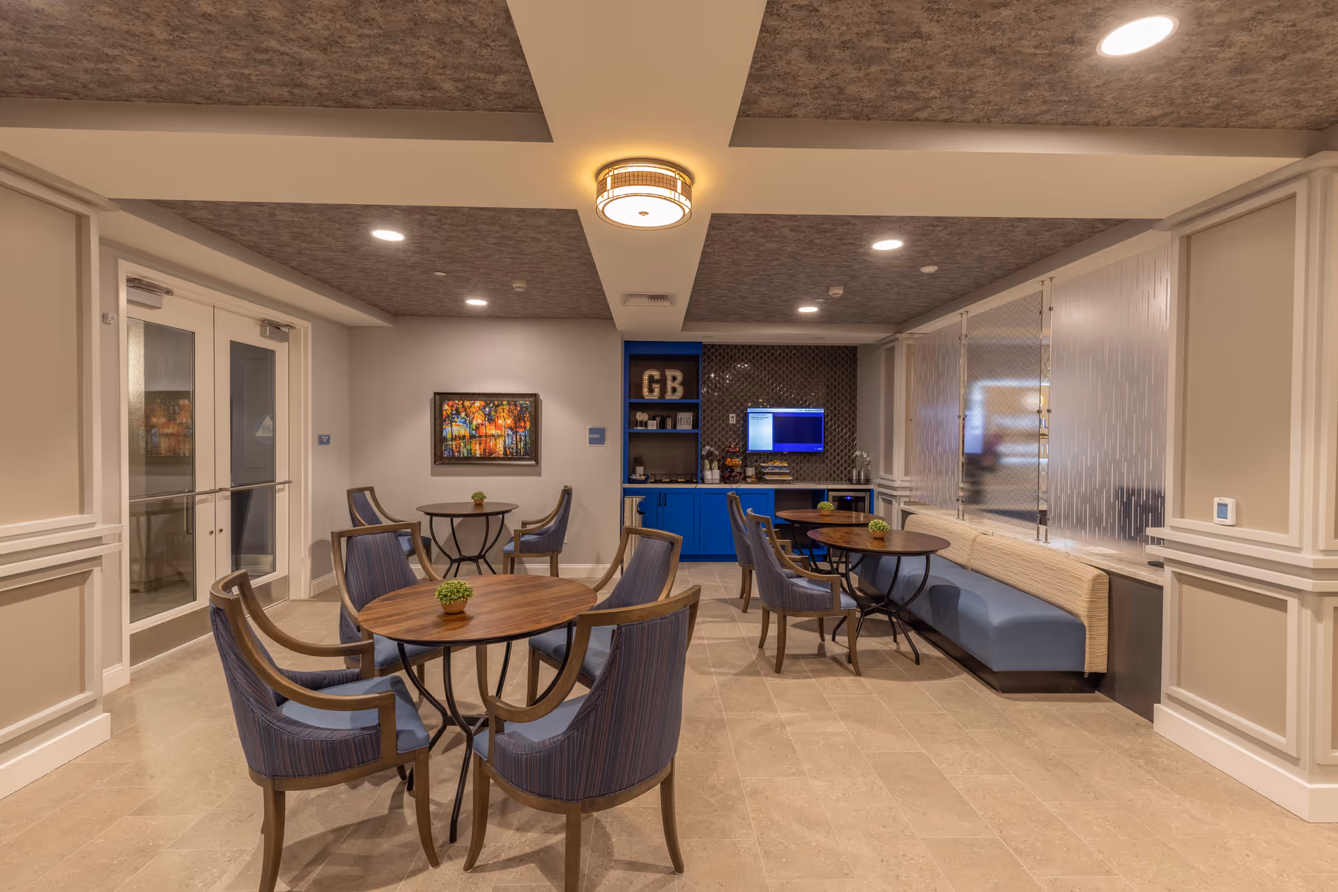 Well-lit communal lounge with round wooden tables, blue upholstered chairs and bench seating facing a small service counter and TV.